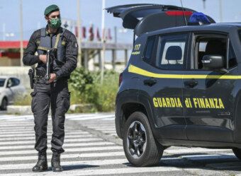 Un agente della Guardia di Finanza in divisa, armato e sorvegliante, accanto a un veicolo di servizio durante un'operazione di sequestro beni