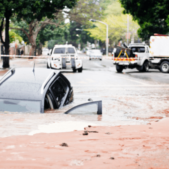 Auto parzialmente sommersa su una strada allagata, con veicoli di soccorso sullo sfondo.